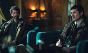 Två män sitter i mörka soffor. Den ena, med skägg och hoodie under jacka, ser fundersam ut. Den andra, med mustasch och kavaj, skriver på ett papper. Kanske ett förhör för dept q.