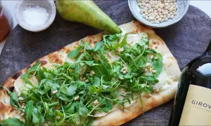 En läcker pinsa med chevre, ruccola och pinjenötter, bredvid en päronhalva, salt och en vinflaska.
