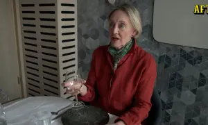 Äldre kvinna med röd skjorta och scarf håller ett glas vin. Hon sitter vid ett dukat bord, redo för en vett och etikett middag.