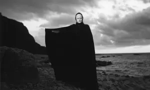 En skrämmande gestalt med vit ansiktsmålning och svart kåpa står på en stenig strand. En ikonisk scen från filmer med Ingmar Bergman.