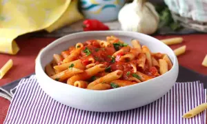 En skål med pasta arrabiata, toppad med färsk persilja. En vitlöksklyfta och en röd chili syns i bakgrunden.