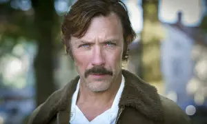 Porträtt av en svensk manlig skådespelare från 2000-talet med mustasch och blå ögon, klädd i en brun jacka med foder.