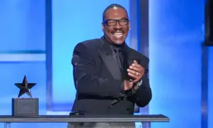 Eddie Murphy, känd för sina roliga filmer, ler och klappar händerna på scenen bredvid en stjärnstatyett.