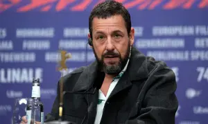 Adam Sandler, känd från många roliga filmer, sitter vid ett bord med en mikrofon och en vattenflaska.