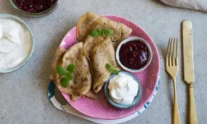 En tallrik med glutenfria pannkakor, serverade med sylt och grädde. En gyllene gaffel och kniv ligger bredvid.