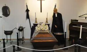En präst med sorgflor på hatten står bredvid en kista med blommor. En orgel och kors syns i bakgrunden.