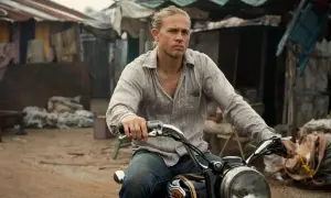 Charlie Hunnam kör motorcykel genom ett slumområde, med en blick av beslutsamhet.