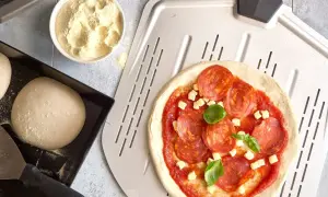 Pizzadeg recept: färdig pizzadeg, tomatsås, salami och ost på en pizzaspade. Bredvid en skål med mjöl och en form med pizzadeg.