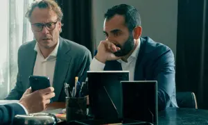 Två män i kostym sitter vid ett bord. Den ena mannen med glasögon tittar på sin mobiltelefon, den andra mannen funderar. Scen från "Hundarna" på SVT.