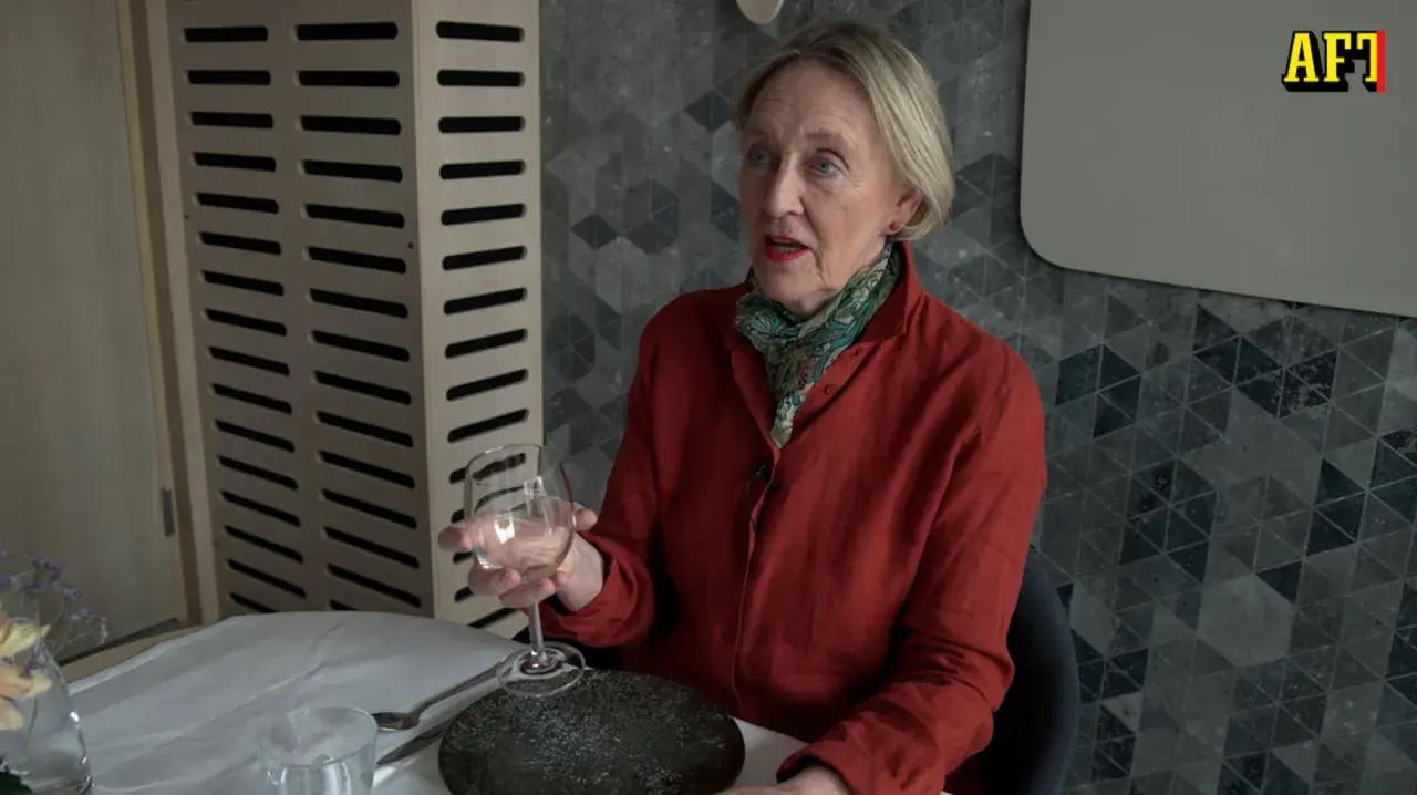 Äldre kvinna med röd skjorta och scarf håller ett glas vin. Hon sitter vid ett dukat bord, redo för en vett och etikett middag.