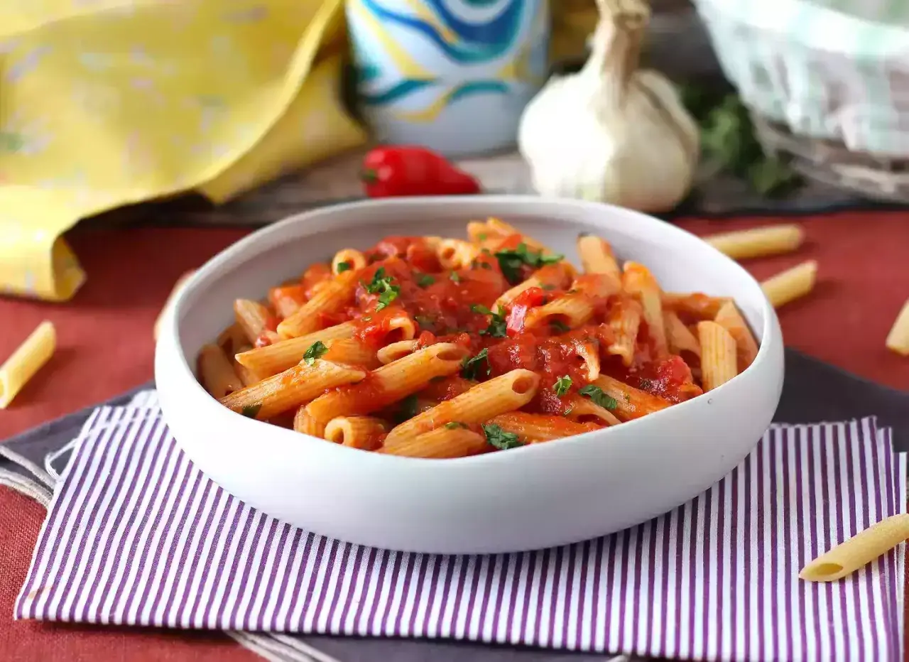 En skål med pasta arrabiata, toppad med färsk persilja. En vitlöksklyfta och en röd chili syns i bakgrunden.