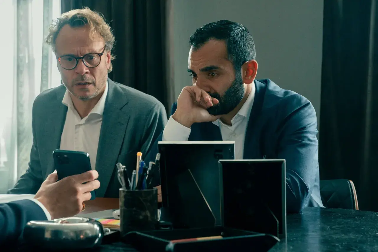 Två män i kostym sitter vid ett bord. Den ena mannen med glasögon tittar på sin mobiltelefon, den andra mannen funderar. Scen från "Hundarna" på SVT.
