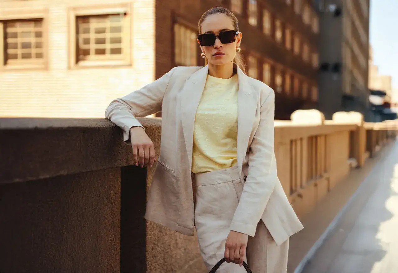 Kvinna i ljusbeige kavaj och byxor, perfekt för business casual dam. Solglasögon och gul topp ger en modern touch.