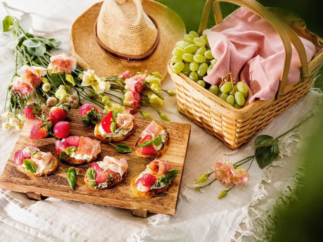 En h&auml;rlig picknick med bruschetta toppad med jordgubbar och prosciutto, f&auml;rska vindruvor i en korg, blommor och en str&aring;hatt. Perfekt picknick mat!