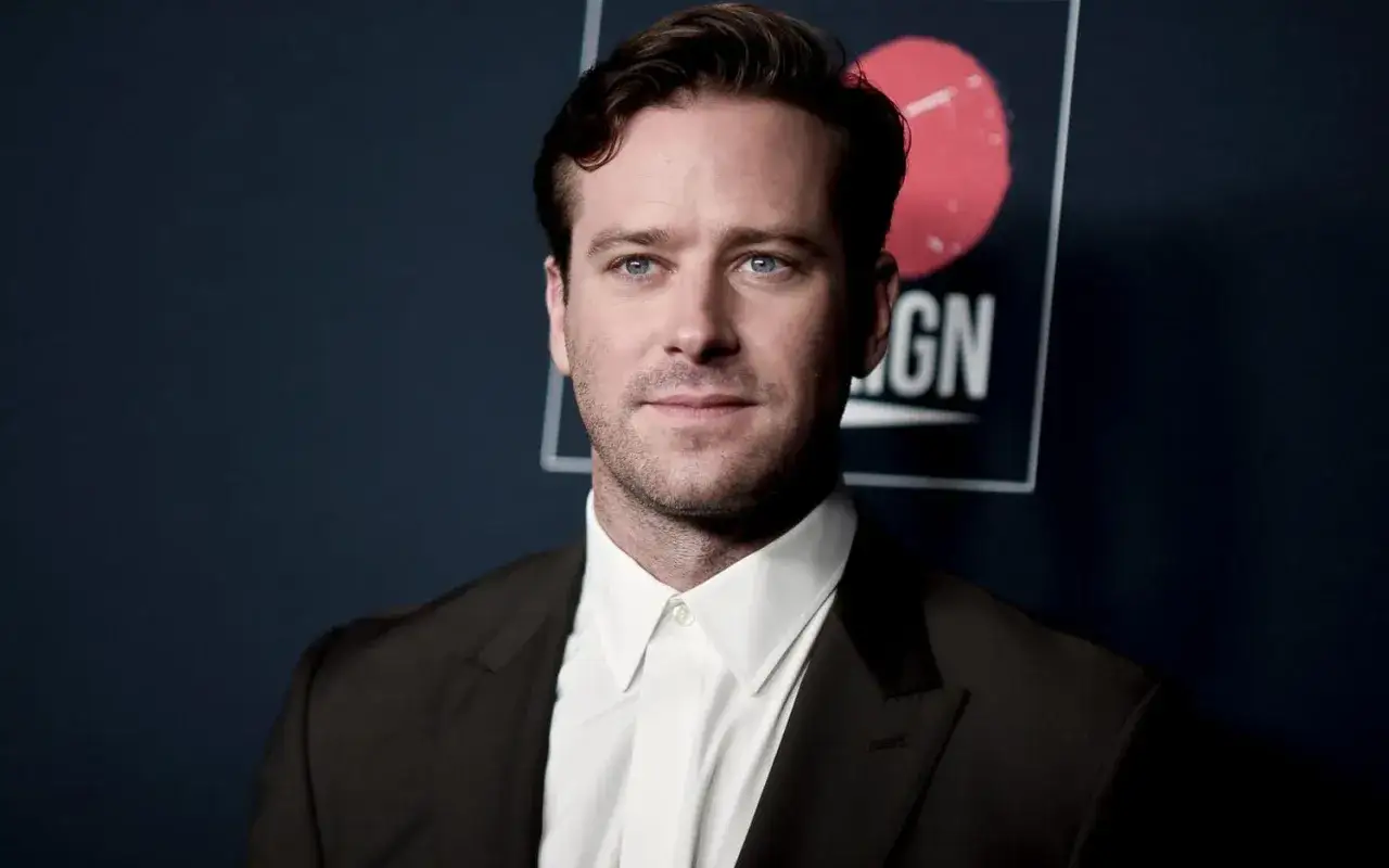 Armie Hammer, kl&auml;dd i svart kostym och vit skjorta, poserar framf&ouml;r en m&ouml;rkbl&aring; bakgrund med en logotyp.