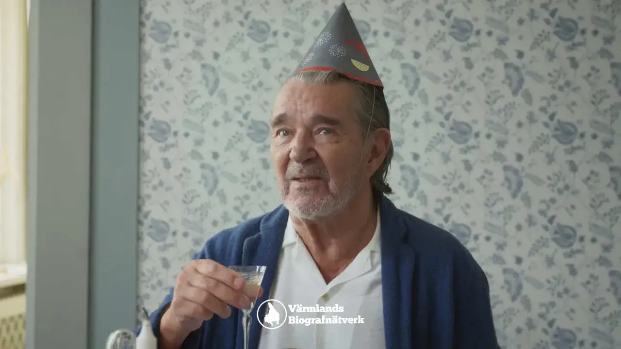 Äldre man med festlig hatt och glas i handen, kanske en scen från grannfejden film.
