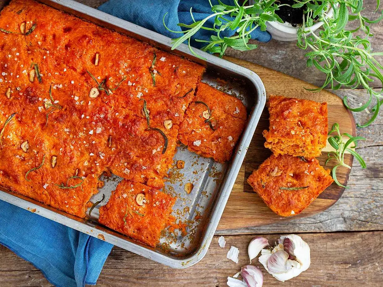 En saftig, orange focaccia med rosmarin och flingsalt, perfekt för ett enkelt focaccia recept. Två bitar ligger på en skärbräda bredvid.