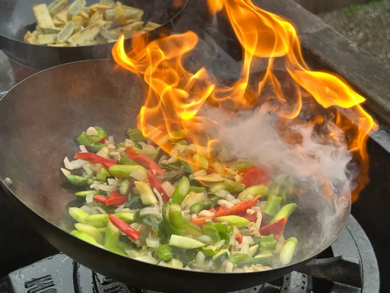 Grönsaker flamberas i en wok, redo för ett snabbt och smakrikt wok recept. I bakgrunden syns friterade potatisstavar.