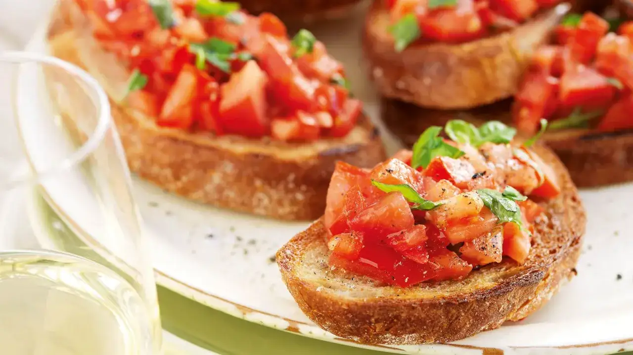 Krispiga bruschettaskivor med f&auml;rska tomater och basilika, perfekt f&ouml;r ett enkelt bruschetta recept. Ett glas vitt vin i f&ouml;rgrunden.