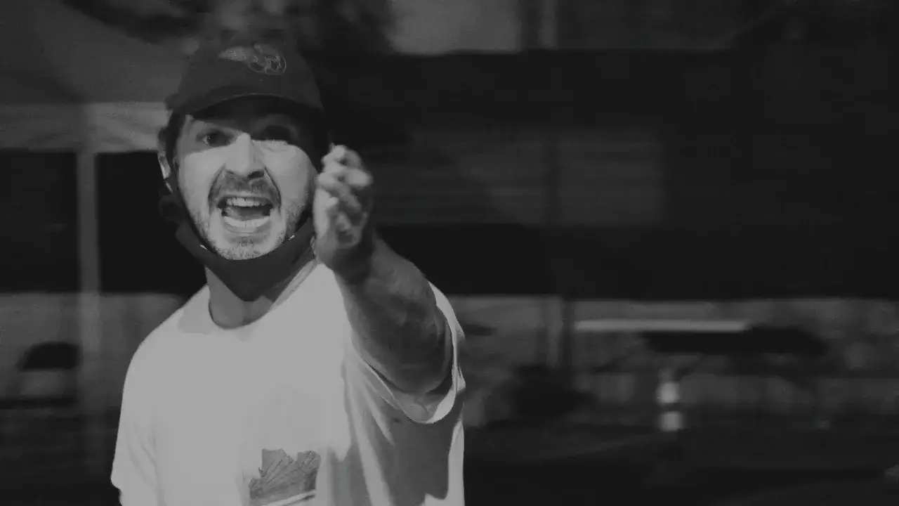 Shia LaBeouf, med keps och munskydd, gestikulerar och ropar med öppen mun.