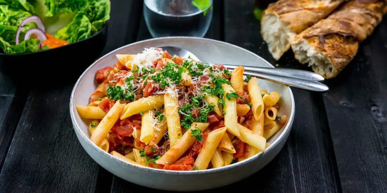 En skål med pasta arrabiata, toppad med persilja och parmesan. Bredvid syns en sallad och ett gott bröd.