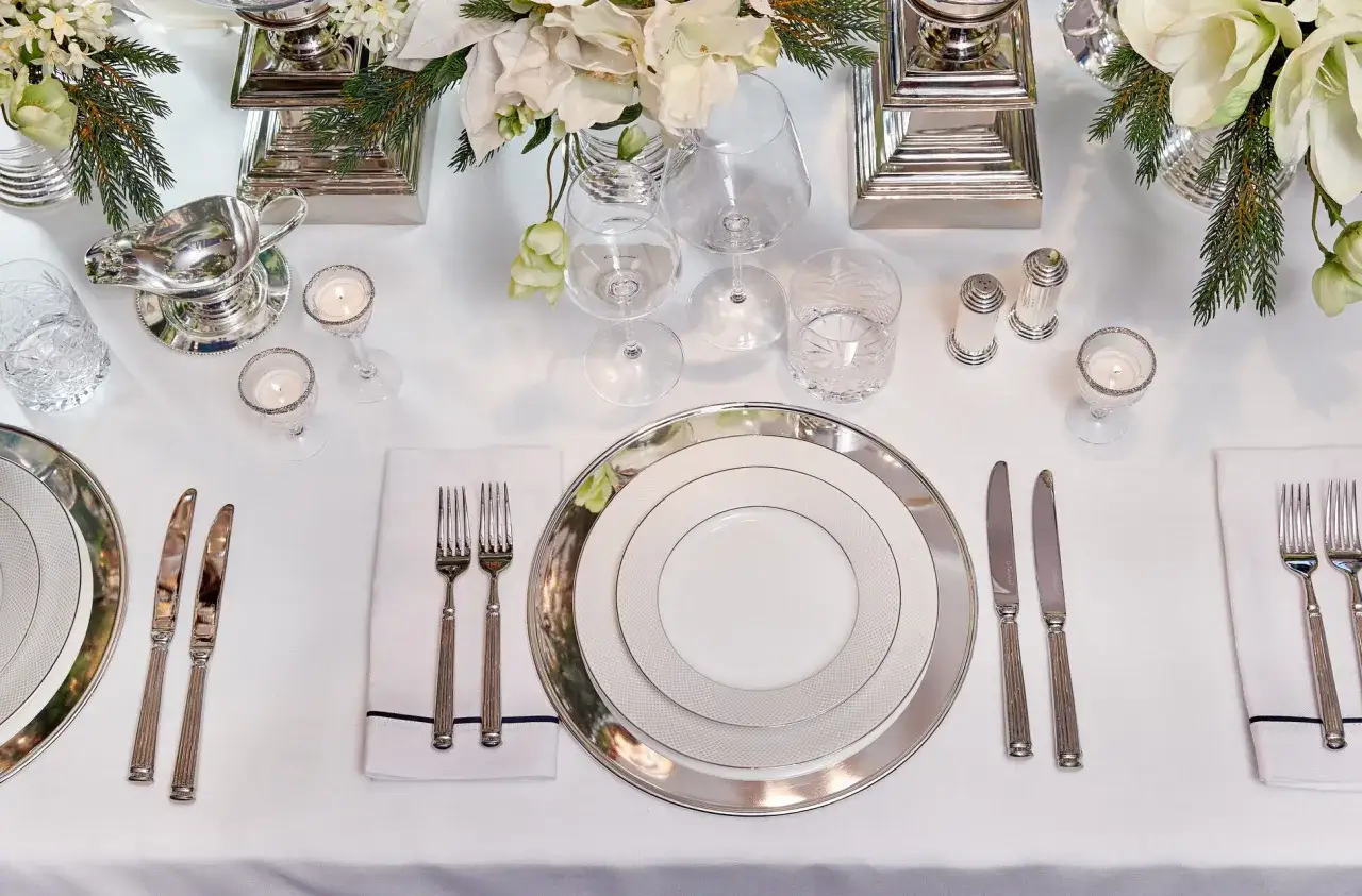 Elegant dukning med silverbestick p&aring; samma sida. Vita blommor och gr&ouml;na kvistar pryder bordet.
