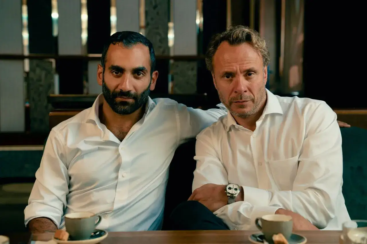Två män i vita skjortor sitter vid ett bord med kaffe. En av dem har armen om den andres axel. Scen från