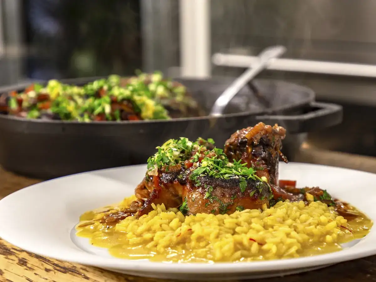 En tallrik med osso buco, saftigt kött med ben, serverat på en bädd av gyllene risotto. I bakgrunden syns en gryta med grönsaker.